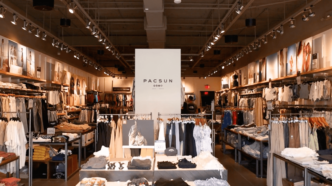 Interior of a PacSun clothing store with racks of clothes, neatly folded items on tables, and a white display wall with the store’s name.